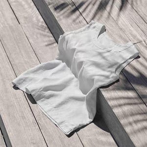 May include: A white linen top with a scoop neck, resting on a wooden surface. The fabric is slightly wrinkled, and the sunlight casts shadows from overhead foliage. The top appears to be sleeveless.