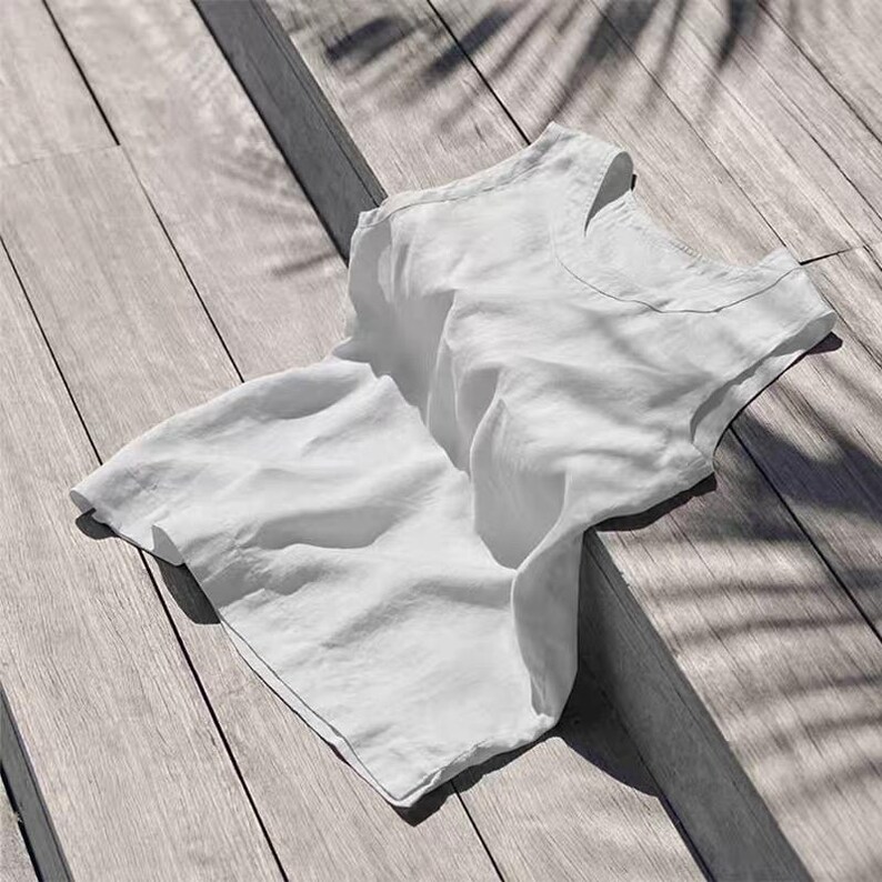 May include: A white linen top with a scoop neck, resting on a wooden surface. The fabric is slightly wrinkled, and the sunlight casts shadows from overhead foliage. The top appears to be sleeveless.