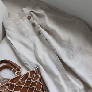 Pu&ograve; includere: Pantaloni in lino beige con chiusura a bottoni. I pantaloni sono piegati su una superficie bianca, con una borsa in pelle marrone con un design a rete in primo piano. I pantaloni hanno una vestibilit&agrave; rilassata e un aspetto naturale.
