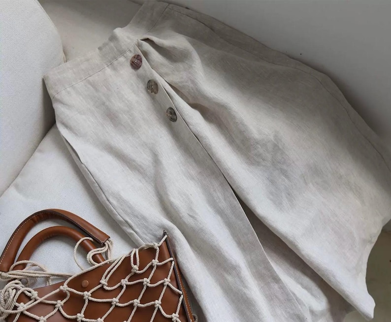 Pu&ograve; includere: Pantaloni in lino beige con chiusura a bottoni. I pantaloni sono piegati su una superficie bianca, con una borsa in pelle marrone con un design a rete in primo piano. I pantaloni hanno una vestibilit&agrave; rilassata e un aspetto naturale.