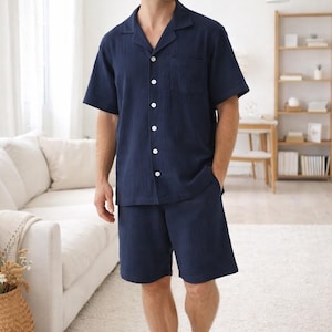 May include: Navy blue pajama set. The set includes a short-sleeved button-down shirt with a chest pocket and matching shorts. The fabric has a textured appearance. The model is standing barefoot on a white rug in a well-lit room.