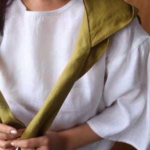 May include: White linen top with elbow-length sleeves and a round neckline. A folded olive green scarf is draped over the shoulder. The scarf is made of a similar linen material. The outfit is simple and elegant.