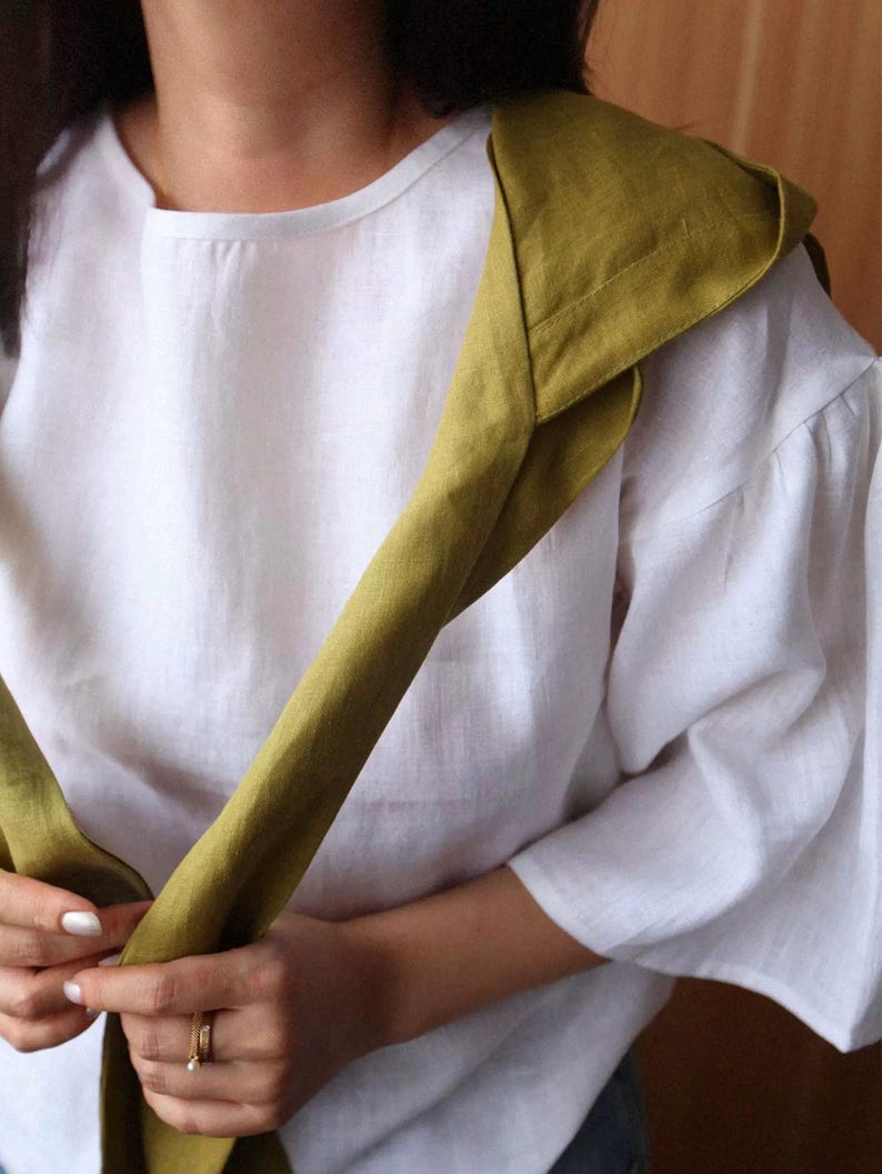 May include: White linen top with elbow-length sleeves and a round neckline. A folded olive green scarf is draped over the shoulder. The scarf is made of a similar linen material. The outfit is simple and elegant.