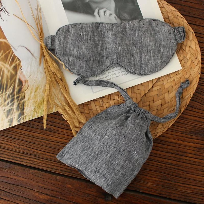 May include: A gray linen sleep mask and matching drawstring pouch. The sleep mask is oval-shaped with a curved nose bridge and ear straps. The pouch is a small, rectangular bag with a drawstring closure. The items are on a woven mat.