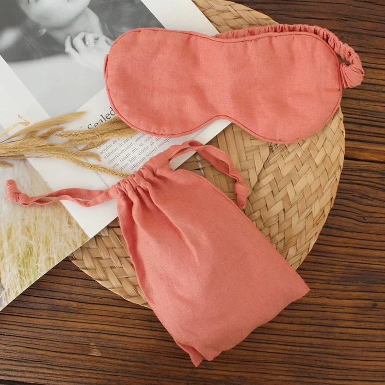May include: A coral-colored sleep mask and matching drawstring pouch. The mask has an elastic band and is made of a soft, textured fabric. The pouch is closed with a drawstring. The items are displayed on a woven mat.