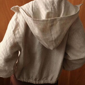 May include: A light grey hooded jacket made of linen. The jacket features a drawstring hem and a large hood. The sleeves are long and the jacket appears to be a relaxed fit. The back of the jacket is visible.