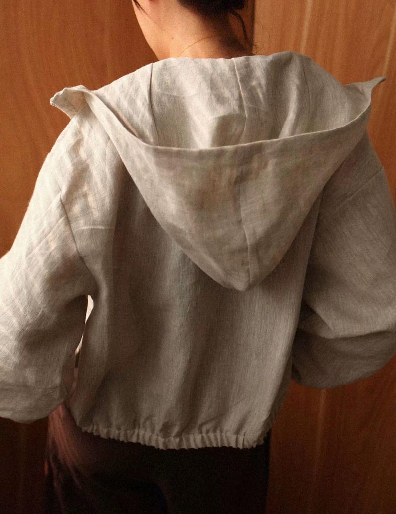May include: A light grey hooded jacket made of linen. The jacket features a drawstring hem and a large hood. The sleeves are long and the jacket appears to be a relaxed fit. The back of the jacket is visible.
