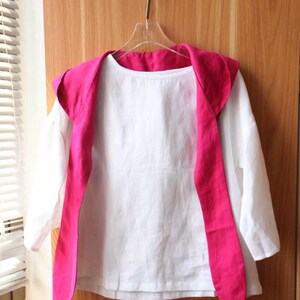 May include: A white long-sleeved shirt with a bright pink shawl collar and matching long ties, hanging on a clear hanger. The shirt is made of a lightweight fabric, and the collar and ties add a pop of color to the simple design.