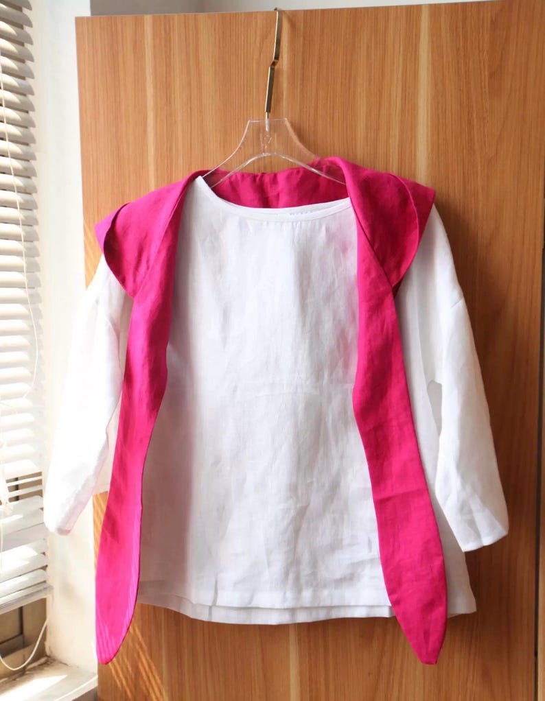May include: A white long-sleeved shirt with a bright pink shawl collar and matching long ties, hanging on a clear hanger. The shirt is made of a lightweight fabric, and the collar and ties add a pop of color to the simple design.