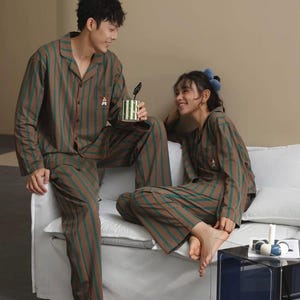 Peut inclure: Ensemble de pyjama à rayures marron et vertes. Le haut à manches longues et le pantalon présentent des rayures verticales. Un petit motif brodé est sur la poche du haut. L'ensemble est porté par deux personnes assises sur un canapé blanc. Une tasse rayée est tenue par une personne.