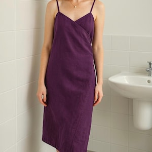 May include: A dark purple linen wrap dress featuring delicate spaghetti straps and a V-neck. The dress, with a knee-length hem, has a wrap design. The fabric looks lightweight and breathable. The setting is a bathroom, with a white sink and tiled wall.