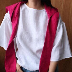 May include: A white linen top with short sleeves and a round neckline. A bright pink scarf is draped over the shoulders and hangs down the front. The outfit is simple and elegant.