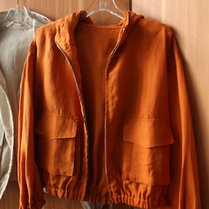 May include: An orange hooded jacket with a full-length zipper and two front pockets. The jacket is made of a lightweight fabric and has a drawstring at the waist. It is hanging on a clear hanger.