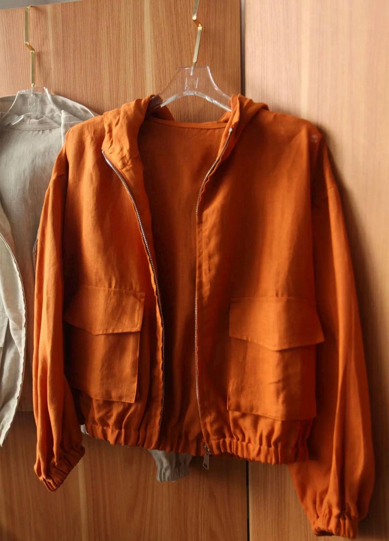 May include: An orange hooded jacket with a full-length zipper and two front pockets. The jacket is made of a lightweight fabric and has a drawstring at the waist. It is hanging on a clear hanger.