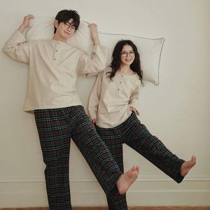 May include: Matching beige pajama sets with long-sleeved tops and plaid trousers. The tops have a Henley neckline. The trousers are a dark plaid with green accents. The people are holding pillows.