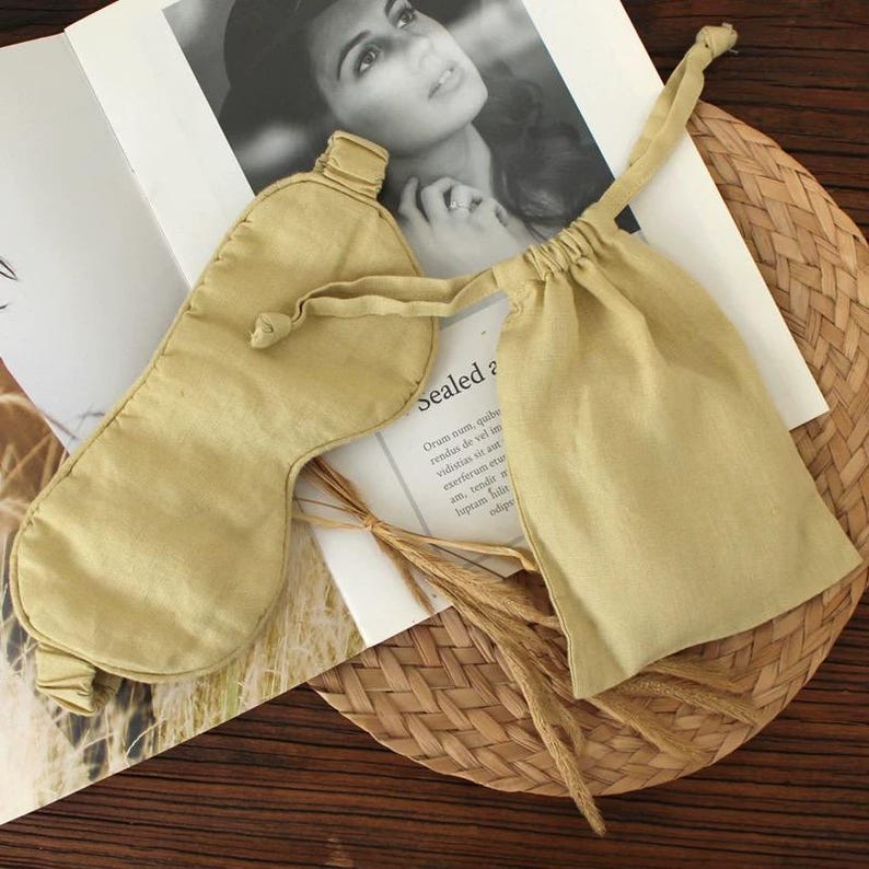 May include: A light tan sleep mask and matching drawstring pouch are displayed on a woven mat. The mask is oval-shaped with a strap, and the pouch is rectangular. The background includes a black and white photo and text that reads "Sealed ?".