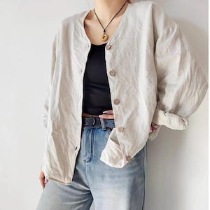 May include: A light beige linen cardigan with wooden buttons. The cardigan is worn over a black top and paired with blue jeans. A black necklace with a circular pendant is also visible.