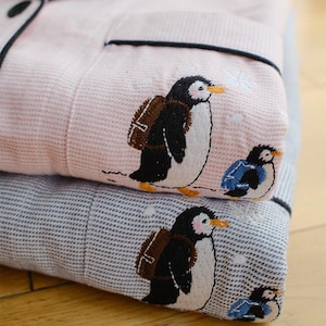 May include: Folded pink and blue pajama set with embroidered penguin designs. The pink pajamas have a striped pattern, while the blue ones have a dotted pattern. Each penguin has a backpack and is walking. The pajamas have black trim.