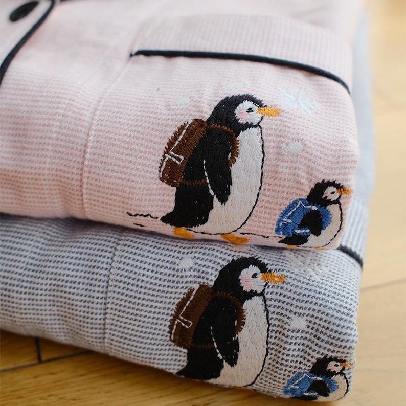 May include: Folded pink and blue pajama set with embroidered penguin designs. The pink pajamas have a striped pattern, while the blue ones have a dotted pattern. Each penguin has a backpack and is walking. The pajamas have black trim.