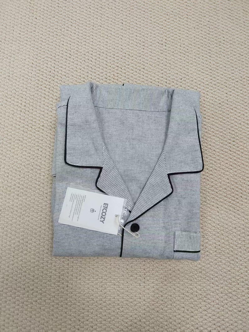 May include: Folded gray pajama set with black trim and a small pocket. The set includes a button-down shirt and pants. A white tag with the text "ETCOZY" is attached to the shirt.