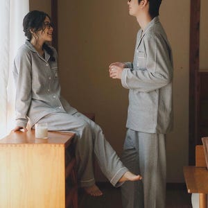 May include: Matching grey and white striped pyjama sets. The long-sleeved button-down shirts and trousers have white trim. A woman sits on a wooden surface, and a man stands nearby. A candle sits on the wooden surface.
