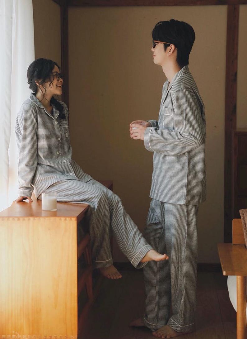 May include: Matching grey and white striped pyjama sets. The long-sleeved button-down shirts and trousers have white trim. A woman sits on a wooden surface, and a man stands nearby. A candle sits on the wooden surface.