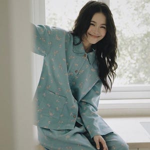 May include: A light blue pajama set featuring a long-sleeved button-down shirt with a collar and a single chest pocket, paired with matching loose-fitting trousers. The fabric is decorated with a repeating pattern of small, light-colored floral motifs.