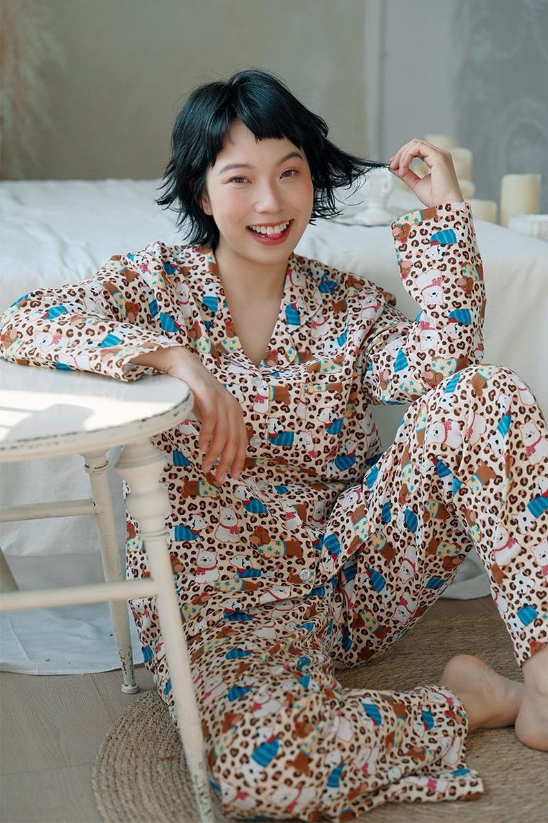 May include: A beige pajama set with a repeating pattern of cartoon bears, blue accents, and leopard print spots. The long-sleeved top has a collar and buttons. The matching pants are also patterned. The person is sitting on a woven mat.