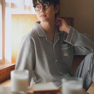 May include: A person wearing a grey and white checked pyjama set with white trim. The pyjamas have a small embroidered design on the pocket. The person is wearing glasses and is sitting near a window. A table with food and drinks is in front of them.