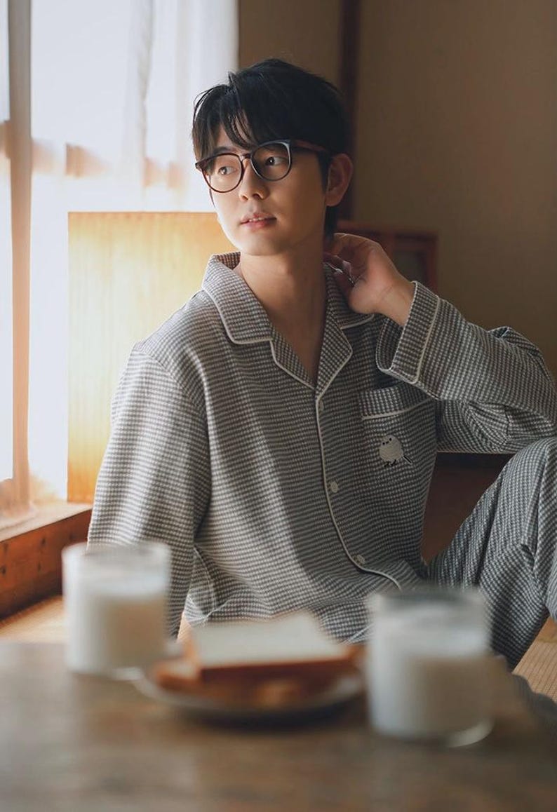 May include: A person wearing a grey and white checked pyjama set with white trim. The pyjamas have a small embroidered design on the pocket. The person is wearing glasses and is sitting near a window. A table with food and drinks is in front of them.