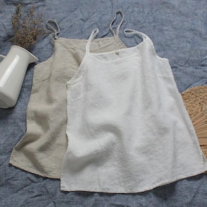 May include: Two linen camisole tops. One is a light beige color, and the other is white. Both tops have thin spaghetti straps and a simple, relaxed fit. They are displayed on a blue fabric surface.