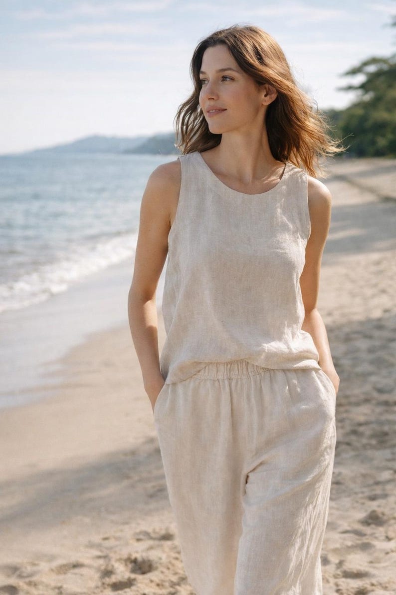 May include: A woman wearing a beige linen sleeveless top and matching pants. The outfit has a relaxed fit and is made of a lightweight fabric. The model is standing on a sandy beach with the ocean in the background.
