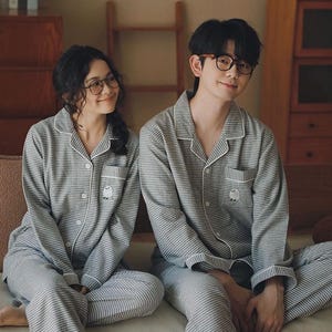 May include: Matching grey and white plaid pyjamas. The long-sleeved button-down shirts and trousers have white trim and a small embroidered owl on the pocket. The couple is wearing glasses and smiling.