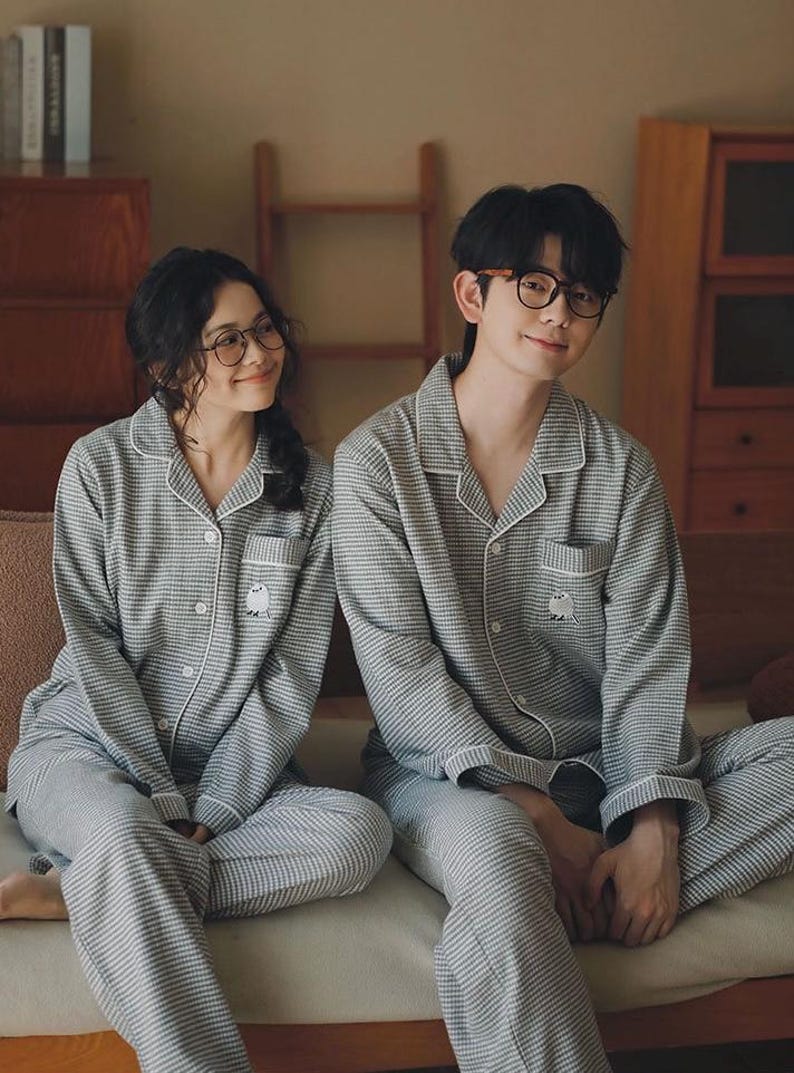 May include: Matching grey and white plaid pyjamas. The long-sleeved button-down shirts and trousers have white trim and a small embroidered owl on the pocket. The couple is wearing glasses and smiling.