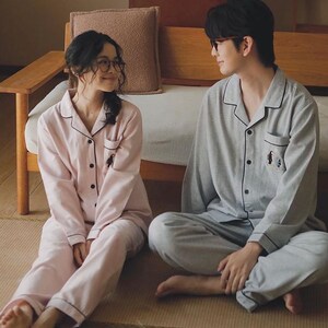 May include: Matching pajama sets in soft pink and gray. The pink set features black trim and a small penguin design on the pocket. The gray set has black trim and a penguin design. Both sets include a button-down top and pants. The couple is sitting on a woven mat.