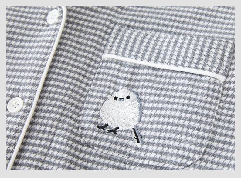 May include: Close-up of a grey and white houndstooth pajama top with white piping and buttons. A small pocket features an embroidered white bird with black details. The pajama top is made of a soft, textured fabric.