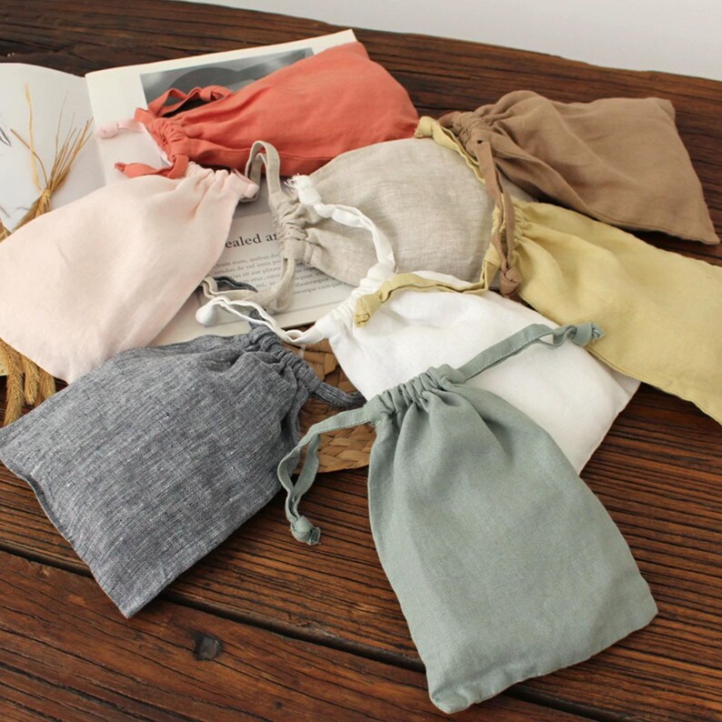May include: A collection of small, drawstring linen bags in various colors, including pink, gray, white, and tan. The bags are arranged on a wooden surface, with some overlapping. The bags appear to be empty and ready for use.