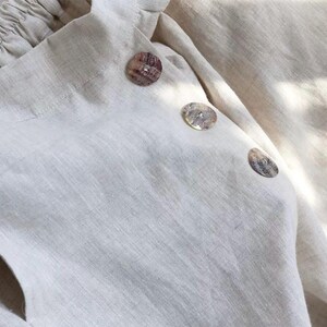 Pu&ograve; includere: Primo piano di un capo in lino beige chiaro con tre bottoni decorativi a conchiglia. Il tessuto ha un aspetto strutturato e i bottoni sono rotondi con una finitura naturale e iridescente. Il capo ha uno scollo arricciato.