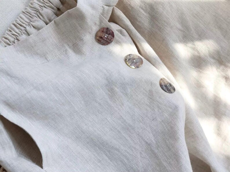 Pu&ograve; includere: Primo piano di un capo in lino beige chiaro con tre bottoni decorativi a conchiglia. Il tessuto ha un aspetto strutturato e i bottoni sono rotondi con una finitura naturale e iridescente. Il capo ha uno scollo arricciato.