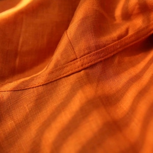 May include: Close-up of orange fabric with visible texture and a stitched seam. The fabric appears to be linen or a similar woven material, with soft folds and subtle variations in color.