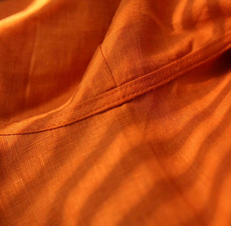 May include: Close-up of orange fabric with visible texture and a stitched seam. The fabric appears to be linen or a similar woven material, with soft folds and subtle variations in color.