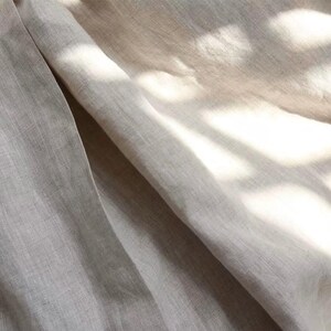 Pu&ograve; includere: Primo piano di un tessuto di lino beige chiaro con trama e pieghe visibili. Il tessuto &egrave; illuminato dalla luce solare, creando ombre e riflessi. L'immagine mostra l'aspetto naturale e organico del tessuto, adatto per abbigliamento o arredamento.