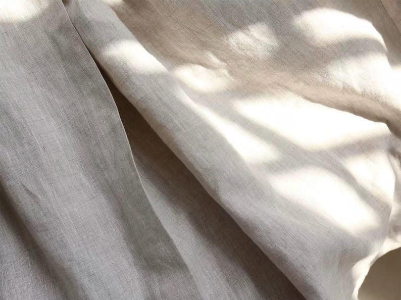 Pu&ograve; includere: Primo piano di un tessuto di lino beige chiaro con trama e pieghe visibili. Il tessuto &egrave; illuminato dalla luce solare, creando ombre e riflessi. L'immagine mostra l'aspetto naturale e organico del tessuto, adatto per abbigliamento o arredamento.