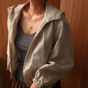 May include: A light beige hooded jacket with a zipper closure and a pocket on the front. The jacket is made of a lightweight fabric and has elastic cuffs. A gray tank top is visible underneath.