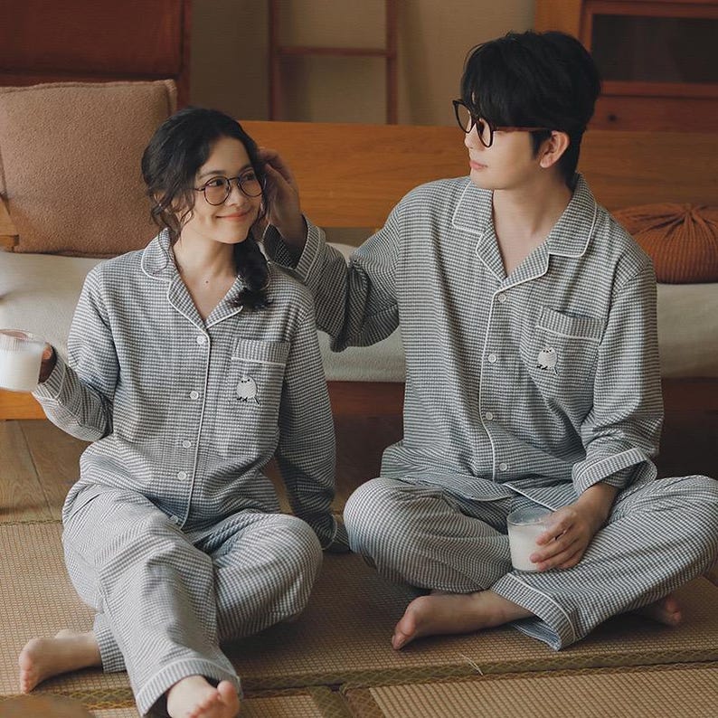 May include: Matching grey and white striped pyjama sets. Each set includes a button-down shirt with a small pocket and a pair of trousers. The shirts have a small embroidered design on the pocket. The pyjamas are worn by two people.