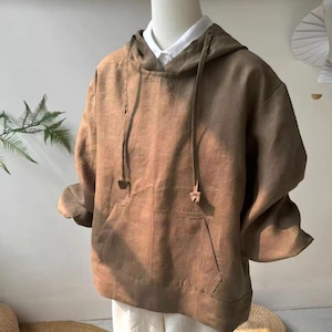 Puede incluir: Una sudadera con capucha marrón con un bolsillo delantero y cordón. La sudadera está hecha de una tela suave y texturizada y se muestra en un maniquí. Debajo se ve una camisa blanca con cuello.