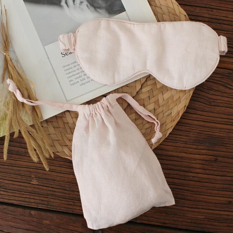 May include: A light pink sleep mask and matching drawstring pouch. The mask is oval-shaped with a strap, and the pouch is gathered at the top. Both items are made of a soft, textured fabric, and are resting on a woven mat.