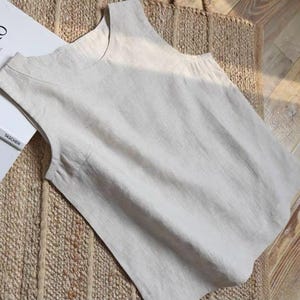 May include: A sleeveless, light beige linen top with a rounded neckline. The top is lying on a woven mat and wooden floor. A book titled "ROTHKO" is visible in the background.