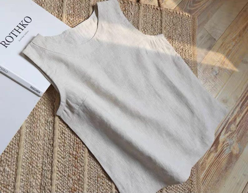 May include: A sleeveless, light beige linen top with a rounded neckline. The top is lying on a woven mat and wooden floor. A book titled "ROTHKO" is visible in the background.
