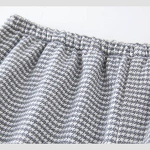 May include: Close-up of grey and white houndstooth fabric, possibly from trousers or shorts. An elasticated waistband is visible at the top, with the fabric gathered below. The material has a soft, textured appearance.